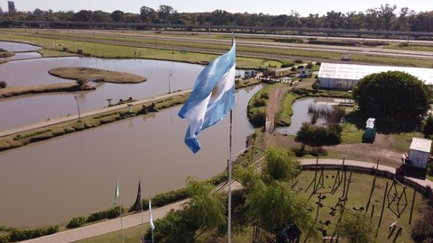 A dynamic high-angle aerial footage of a raised Argentinian flag while waving on a windy day. The pole is surrounded by near establishments, trees, and buildings of the city at the background.
