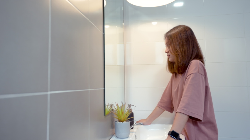 4K, medium shot,  A young Asian woman with long hair is standing in the mirror looking at herself in the bathroom at home. Rub his hands over his hair and face to see details.
