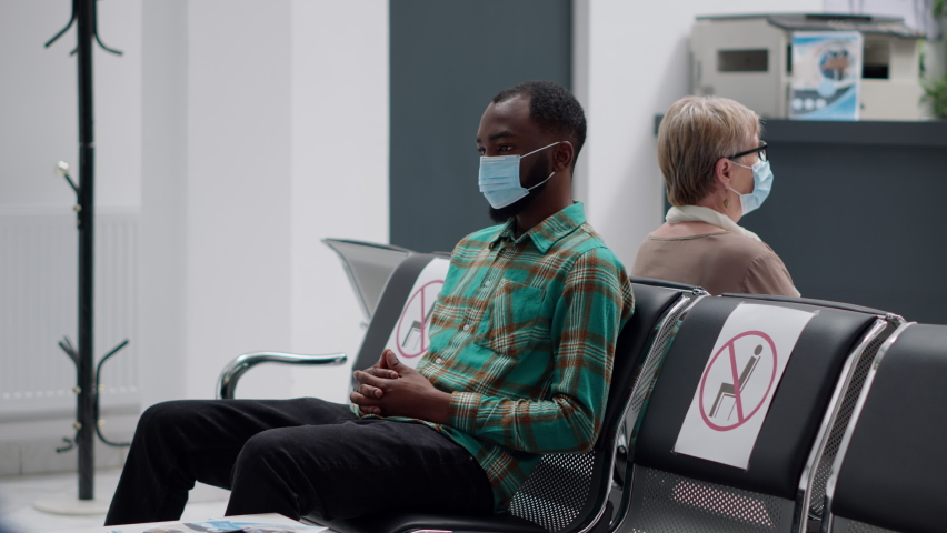 Young man waiting in hospital reception lobby to start consultation with medic during coronavirus pandemic. Diverse people sitting in waiting area chairs to do covid 19 consultation. | Shutterstock HD Video #1092080965