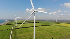 Wind power plant. Windmills with huge propellers. Beautiful landscape with blue sky, green and yellow fields, road and sea cost. Sunny summer day. Aerial view 4K video. Renewable energy production