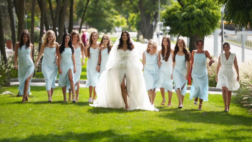 Beautiful bride and her pretty bridemaids in pale blue dresses walking in the park cheering waving hands. Woman with long airy bridal veil in elegant white dress partying with friends. Slow motion. | Shutterstock HD Video #1092083285