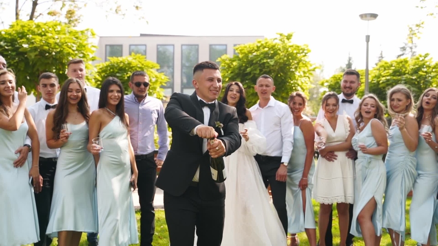 Groom opens champagne bottle splashing wine in the air with excited friends cheering with glasses. Groomsmen and pretty bridesmaids in identical dresses have fun on wedding day in park. Slow motion. | Shutterstock HD Video #1092083287