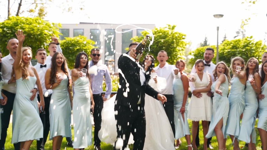 Groom opens champagne bottle splashing wine in the air with excited friends cheering with glasses. Groomsmen and pretty bridesmaids in identical dresses have fun on wedding day in park. Slow motion. | Shutterstock HD Video #1092083287