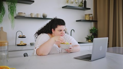 Young plus-size brunette watches movie in cozy kitchen. Curly-haired woman smiles and laughs eating unhealthy crisps. Chubby lady enjoys life staying home