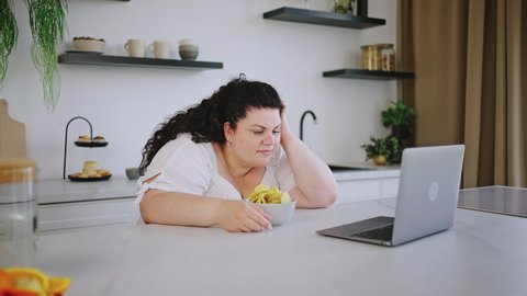 Young overweight curly-haired woman watches movie on laptop eating unhealthy crisps at table. Body positive brunette enjoys life smiling in cozy white kitchen