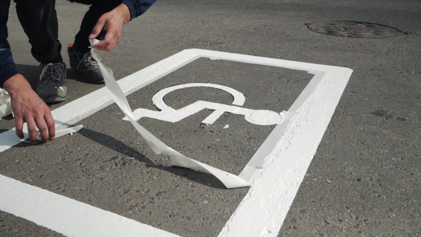 Close-up of manual utility male worker peeling off white tape from freshly painted white disabled car parking lot sign, disability symbol, wheelchair painting on road asphalt surface, summer daytime.