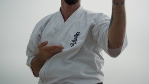 Sporty bearded man exercising karate on cloudy beach close up. Unknown fit fighter training martial art outdoors wearing kimono with black belt. Athletic sportsman honing fight techniques gloomy day.