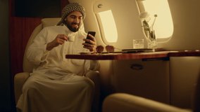 Happy man looking cellphone in private jet. Smiling arabian rest drinking coffee in luxury airplane. Cheerful businessman text smartphone enjoy social media in muslim clothing. Wealthy people concept - Powered by Shutterstock - Get 15% off with code: PIKWIZARD15