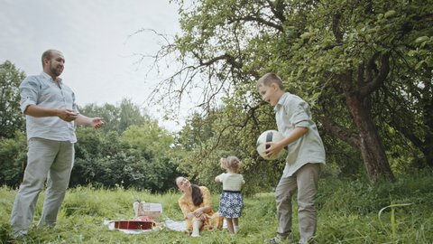 Father and son playing with ball during picnic with family. Low angle tracking shot of happy dad and son throwing and kicking ball to each other near mother resting on blanket and daughter exploring