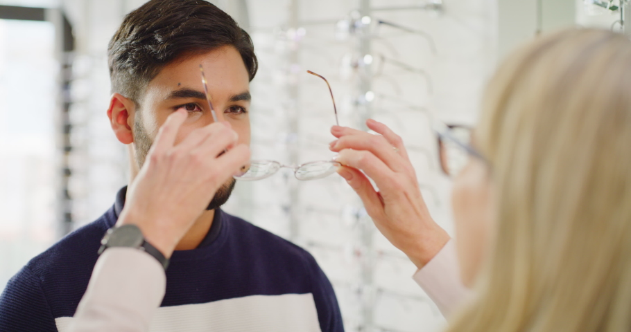 Optician helping a client choose glasses during his optometry appointment. Male customer in consult with optometrist, fitting on a pair of eyewear spectacles for vision corrections or better eyesight Royalty-Free Stock Footage #1092092705