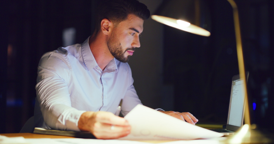Young business man writing and planning his schedule in a diary with a laptop while working late in an office alone. One serious male professional reading documents and brainstorming in a book | Shutterstock HD Video #1092092717