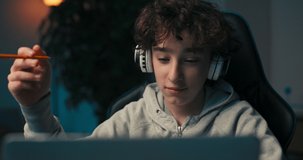A boy with curly hair sits in a room at a desk with a laptop on wireless headphones, taking notes, redrawing, looking off to the side wondering.