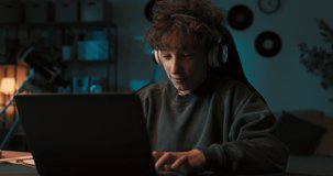 A smiling boy with curly hair wearing wireless headphones works on a school project in front of laptop late into the evening. He listens to music and does his homework.