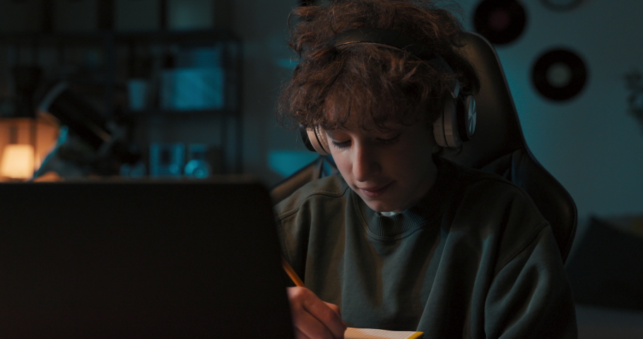 A portrait of a smiling student in remote learning. A young boy sits in a room at a desk with a laptop wearing wireless headphones, taking notes, redrawing.