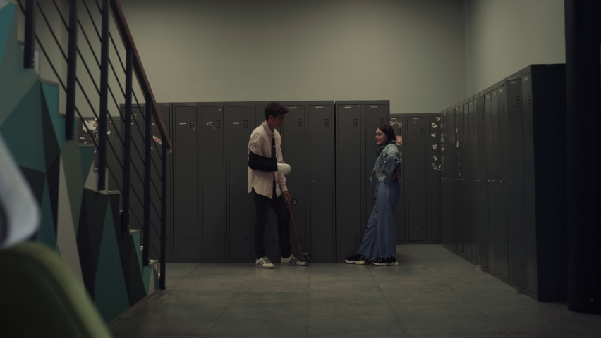 Two students standing talking after classes. Multiethnic couple teens communicating near lockers. Dark-haired indian boy with broken arm chatting with cute girl in empty school hall. Teenagers concept
