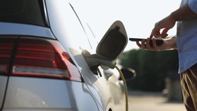 Unrecognizable man plugging electric car from charging station. Male plugging in power cord to electric car using app on smartphone. Businessman charging electric car at outdoor charging station - Powered by Shutterstock - Get 15% off with code: PIKWIZARD15