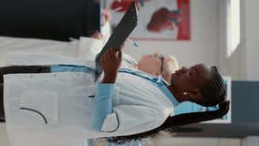 Vertical video: Portrait of african american medic with uniform and checkup report to do consultation in hospital reception. Female physician with healthcare expertise, working in medical system