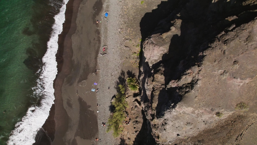 Overhead view of people resting on dark sandy beach with huge rock and cliffs. Aerial static top view of Playa de Guigui beach in the Gran Canaria island. Beautiful sea waves at sunny day in summer.  Royalty-Free Stock Footage #1092111895
