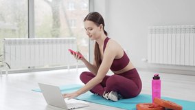 a woman in a burgundy tracksuit with laptop sends a message to students for an online workout a mat in a gym. Online lessons for sports. clothes and shoes for gymnastics. professional trainer. fitness