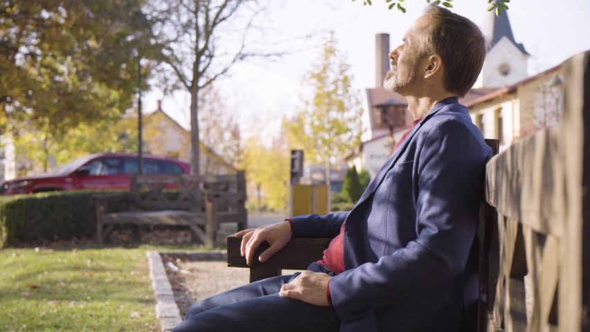 A middle-aged handsome Caucasian man pulls out a smartphone, works on it, then puts it away as he sits on a bench in a quaint town in fall