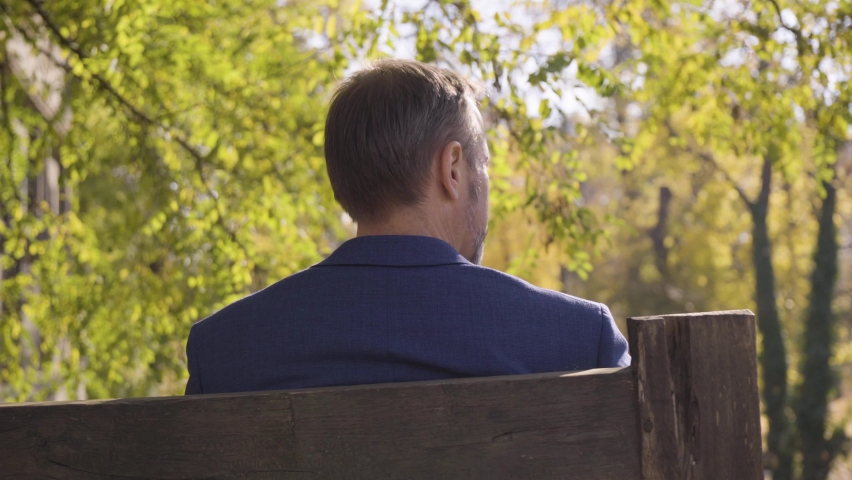 A middle-aged handsome Caucasian man looks around as he sits on a bench in a quaint town in fall - rear closeup