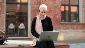 Pretty smiling muslim business woman in hijab sitting on bench and using laptop during video chat. Happy arabic girl sitting outdoor near her work office and checking email on digital gadget.