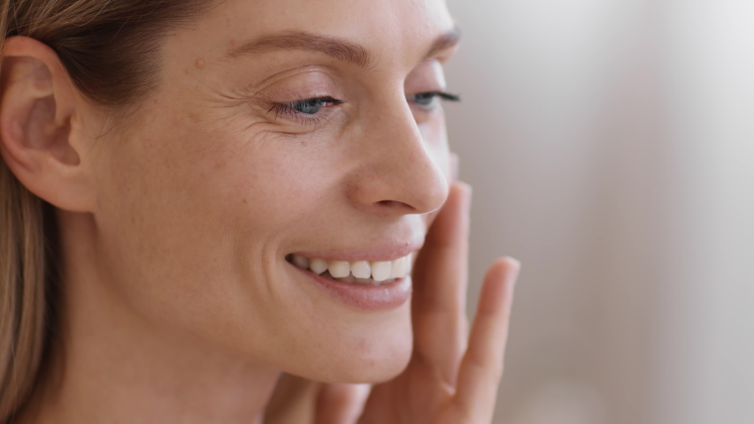 Anti aging skin care. Close up portrait of beautiful middle aged woman with wrinkles applying caring cream on face, touching her skin, tracking shot, slow motion, empty space