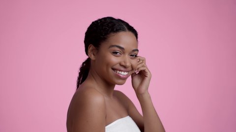 Black beauty concept. Studio portrait of young well-groomed african american woman wrapped in towel touching her smooth face and smiling to camera, pink background, slow motion Arkistovideo