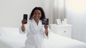 Young happy pregnant lady video chatting with friends via cellphone, showing sonogram and talking to phone, sitting at bathrobe at home, slow motion