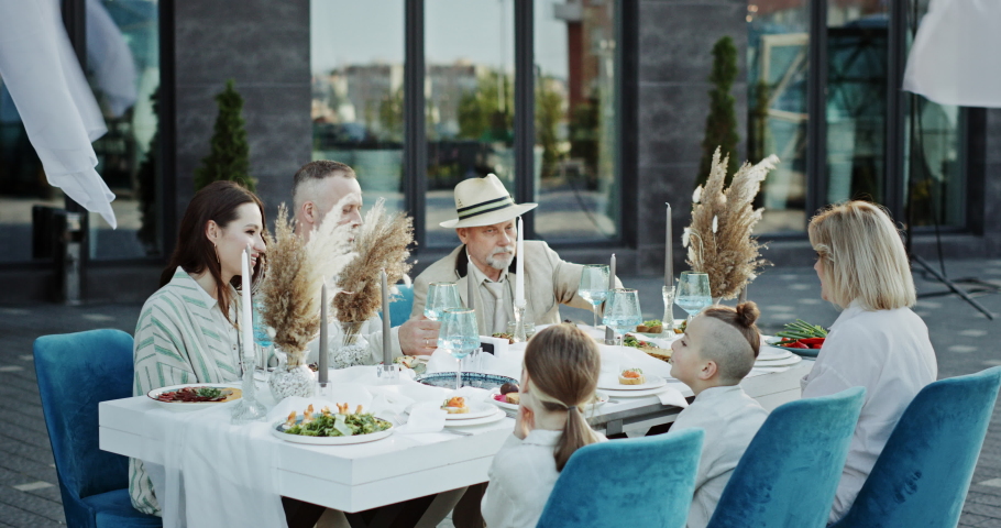 Happy family clinking glasses during banquet in outdoor restaurant. Positive multi generational family in elegant clothes gathering around table and toasting with wineglasses during festive dinner in | Shutterstock HD Video #1092146297