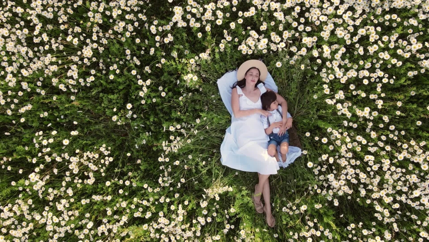 beautiful woman mom in a white dress and hat with a boy son lying on her back in a field with daisies, at sunset in summer. aerial view