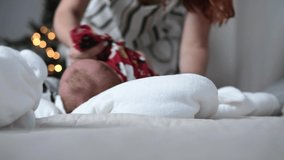 Young mother puts clothes on a newborn baby who lies on the bed in the bedroom at home. In the background is a Christmas tree with a garland. Video