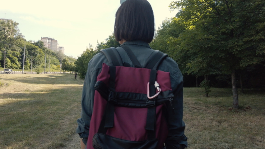 View from behid young woman backpacker walking at the park along the road with red backback. Female traveler at the sunny day