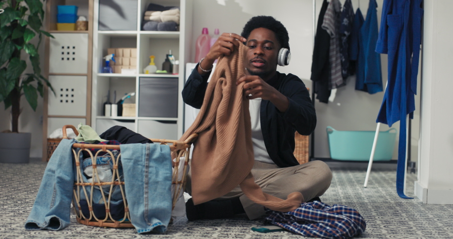 Portrait of a smiling young men folding clean clothes, sorting laundry before putting it in the washing machine, a student sitting on the bathroom floor listening to music on wireless headphones.