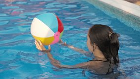 A young girl in a bright leopard swimsuit, swims with a bright colored inflatable ball in a deep blue pool with clear transparent water on a warm sunny summer evening. HD slow-mo video