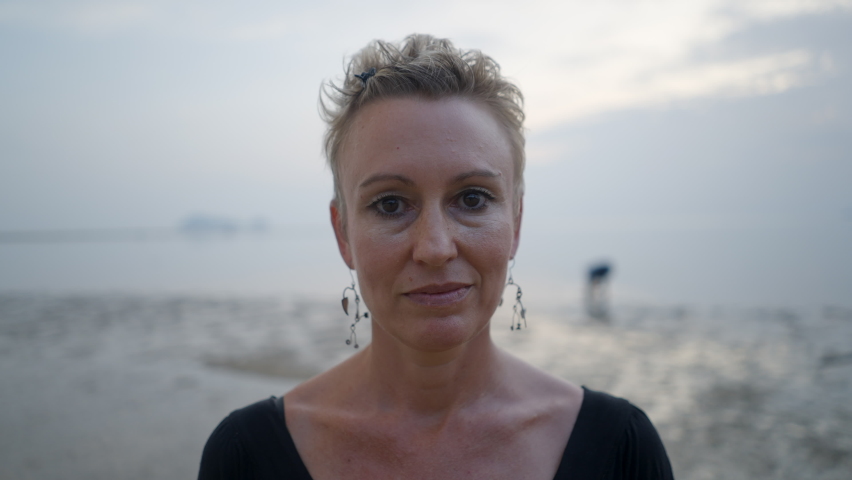 Middle-aged caucasian woman with short blond hair smiling standing at the beach at sunset - bust shot 