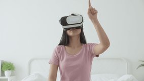 Young asian woman wearing vr headset playing game with excited on bed in the bedroom at home, female using virtual reality or metaverse innovation for simulation 3D, lifestyles and technology concept.