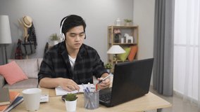 hardworking Chinese young man wearing earphones is writing in notebook and practicing speaking while taking language online course with laptop at home.