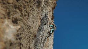 bottom view of muscular strong man rock climber climbs on vertical cliff on rock wall on blue sky background. man puts quickdraw and clips a rope to make belay and be in safe