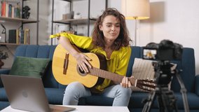 Professional blogger holding music instrument and talking on camera while sitting on comfy sofa. Female guitar performer recording online lesson for vlog using laptop and camera on tripod.