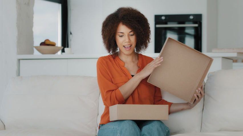 Curious African American buyer customer woman with curly hair sitting at couch at home unbox opening unpacking parcel box gift present purchase order from online shop shipping delivery service indoors | Shutterstock HD Video #1092173333