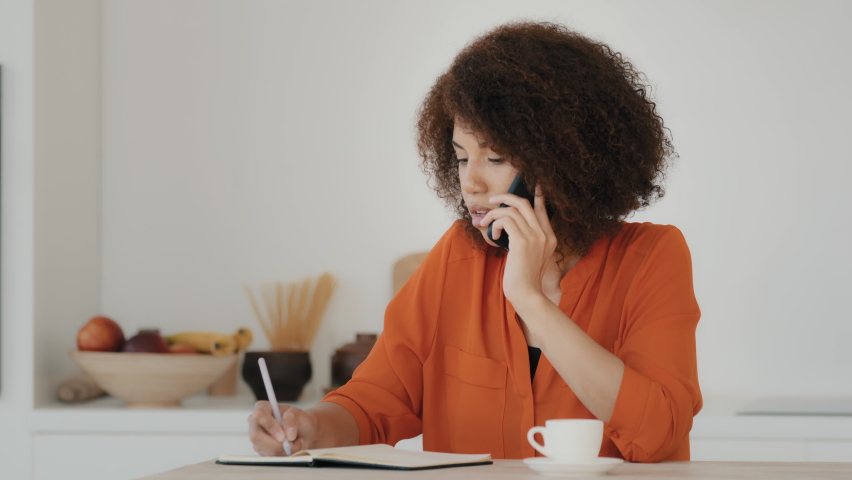 Busy African American business woman curly hair girl student worker housewife working paperwork talking on phone at home kitchen. Ethnic female making order booking call on mobile writing tasks notes Royalty-Free Stock Footage #1092173341