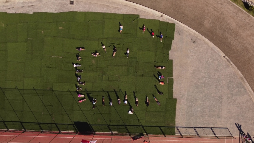 A dynamic descending aerial footage of a group of students lying on the grass of the athletic stadium while doing leg raise exercises.