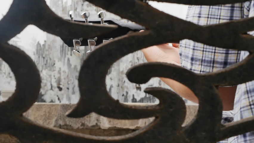 The boy plays the acoustic guitar near the water fountain in the park while sitting on a bench. Rear view.
