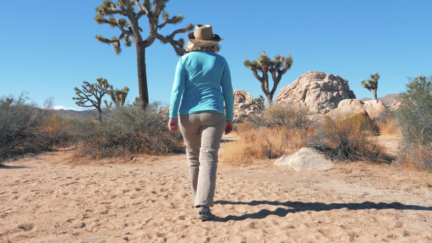 Overweight elderly mature woman walks through sandy arid desert in sunny weather. Hiking in Joshua Tree Park with cacti and thorns, stone monoliths and hills. Stunning landscapes and enjoying nature