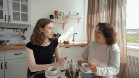 Daughter and mother enjoy drinking tea sitting at glass table in kitchen. Young woman turns to camera to continue talking on video call from third party to POV