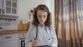 POV of family doctor writing down details listening to patient on video call. Woman with stethoscope sits at kitchen table looking in camera closeup