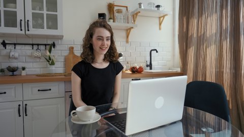 Woman watches movie on laptop getting shocked by scary moment. Young curly-haired lady sits at kitchen table near cup of coffee putting hands on face