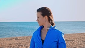 Portrait of beautiful woman looking around and dancing in slow motion by the sea on sandy beach. Her hair flutters in the wind. Happy woman in blue tracksuit against blue sky