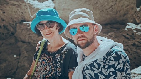 Portrait of Happy senior lady and adult son in sunglasses and panama hats, sitting on rock on mountain hiking trail. Family vacation in warm country. Camera moves from right to left in slow motion